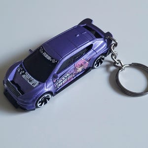 May include: A purple die-cast car keyring with black wheels and a silver key ring. The car features anime-style graphics and text on the side and bonnet. The keyring is a miniature car, possibly a collectible or novelty item.