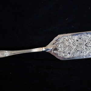 May include: A silver cake server with an ornate, engraved design on the blade. The handle is also silver and has a decorative, scalloped edge.