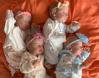 May include: Five realistic baby dolls dressed in various pastel-colored outfits, including onesies and patterned pajamas. Each doll has a headband with a bow or flower. The dolls are lying on an orange fabric.