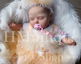 May include: A lifelike baby doll wearing a peach-colored tulle dress and a white headband with a bow. The doll has a pacifier and is nestled in a white basket with fluffy white faux fur. The text "HMunique Etsy Shop" is visible.