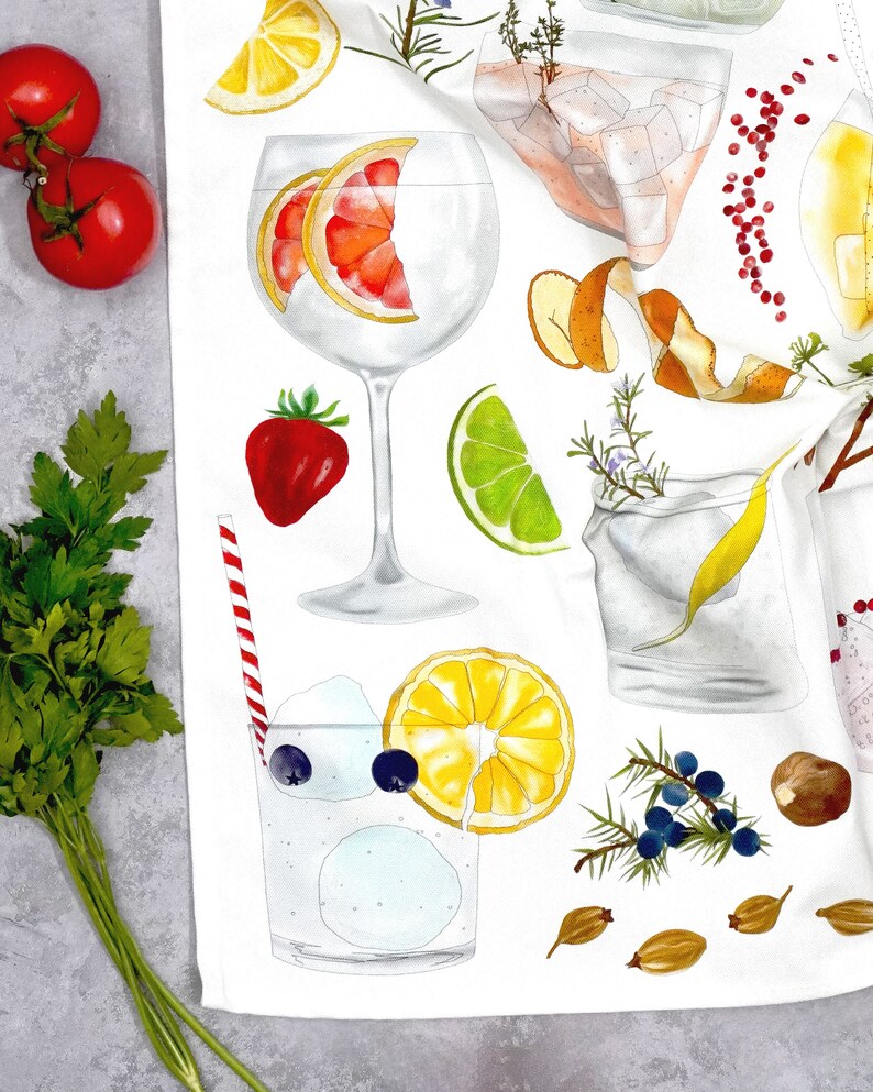 May include: A white kitchen towel with a colorful illustration of various ingredients for a gin and tonic cocktail. The illustration includes a gin glass with a grapefruit slice, a lime wedge, a sprig of rosemary, a lemon wedge, juniper berries, and a sprig of thyme.