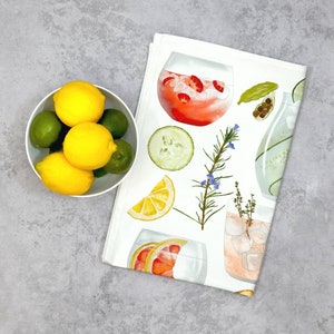 May include: A white tea towel with a colorful illustration of various ingredients for a gin and tonic, including lemons, limes, cucumbers, rosemary, and juniper berries. The towel features a variety of gin and tonic cocktails in glasses.
