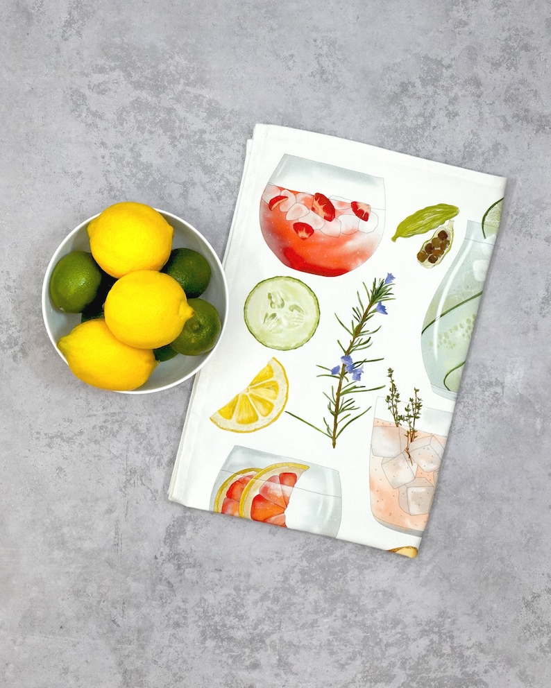 May include: A white tea towel with a colorful illustration of various ingredients for a gin and tonic, including lemons, limes, cucumbers, rosemary, and juniper berries. The towel features a variety of gin and tonic cocktails in glasses.