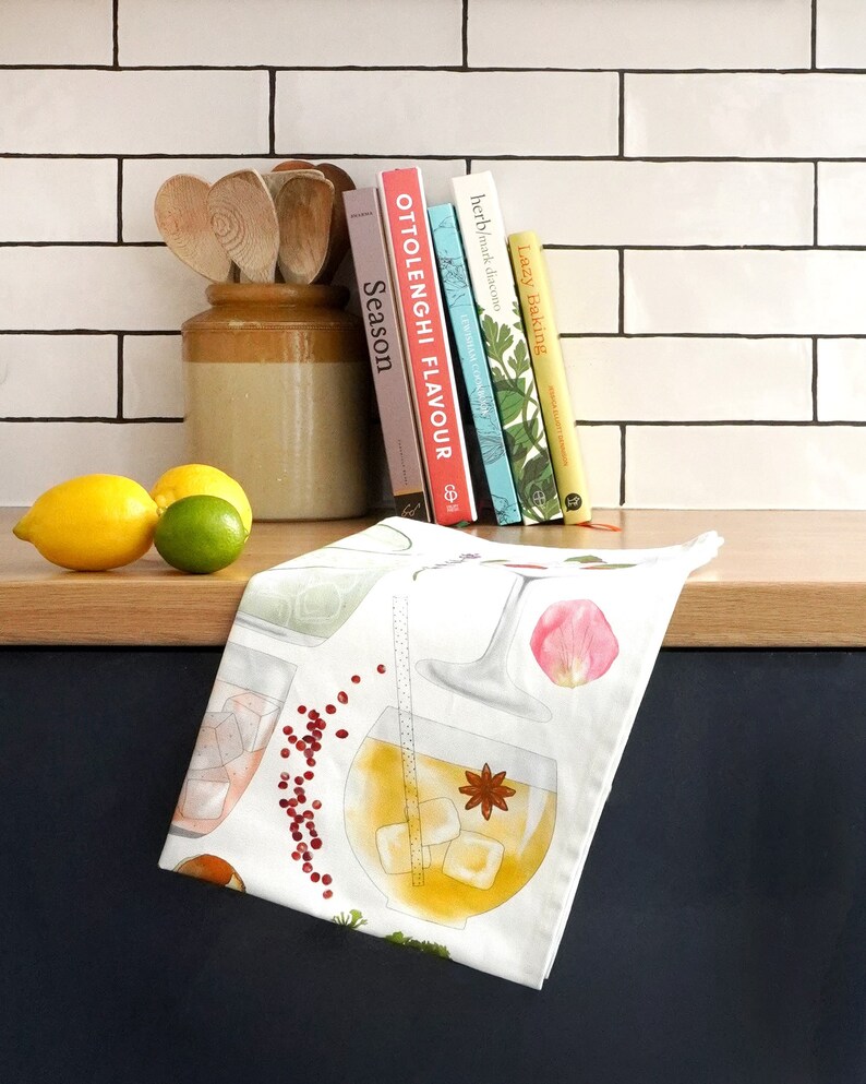 May include: White tea towel with a colorful illustration of cocktails and ingredients. The towel features a variety of drinks, including a gin and tonic, a margarita, and a Moscow mule. The towel is perfect for adding a touch of whimsy to your kitchen.