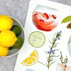 May include: A white kitchen towel with watercolor illustrations of various ingredients for making a gin and tonic. The ingredients include a slice of lemon, cucumber, rosemary sprigs, thyme, and a glass of gin and tonic with ice and strawberries.