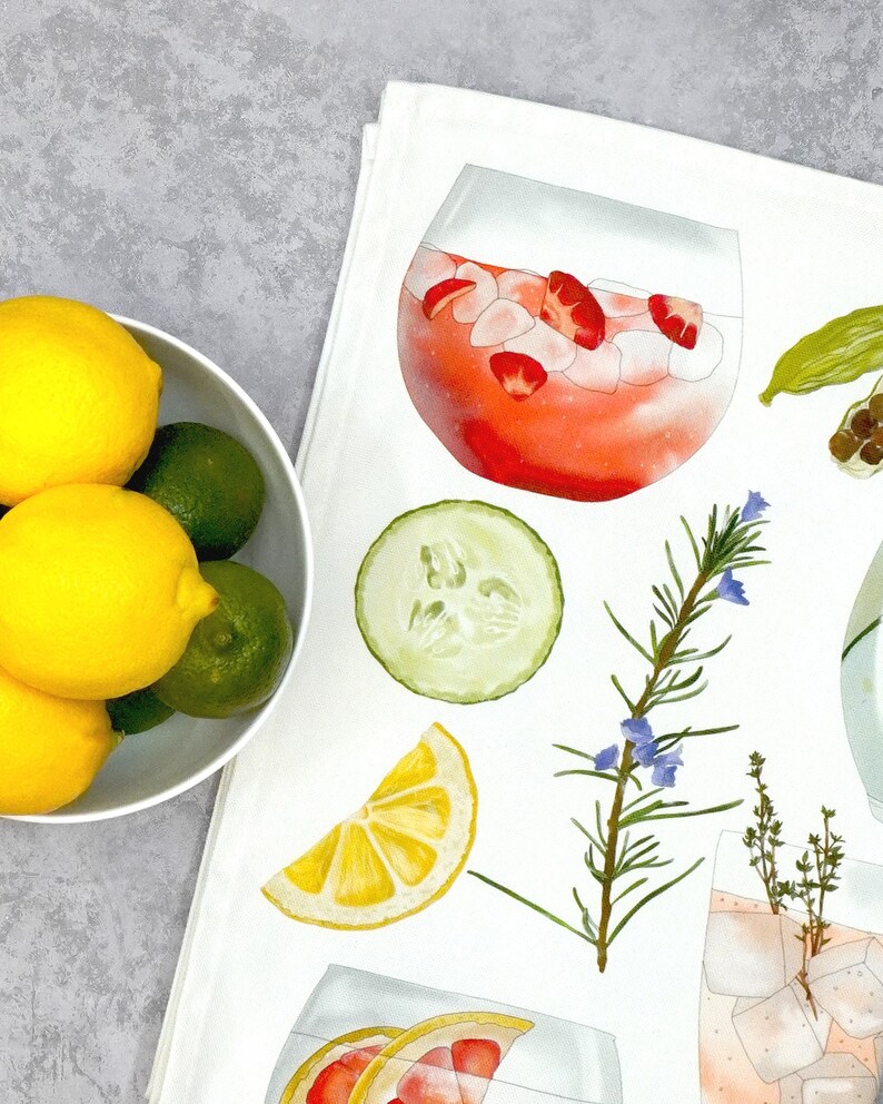 May include: A white kitchen towel with watercolor illustrations of various ingredients for making a gin and tonic. The ingredients include a slice of lemon, cucumber, rosemary sprigs, thyme, and a glass of gin and tonic with ice and strawberries.