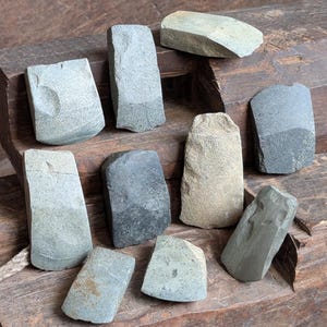 May include: A collection of ten hand-shaped stone tools in various shades of gray and tan. The tools are arranged on a weathered wooden surface, showcasing their unique shapes and textures. The tools vary in size and shape.