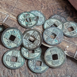 Peut inclure: Un ensemble de pièces de monnaie chinoises antiques avec des trous carrés au centre, disposées sur une surface en bois vieilli. Les pièces présentent une patine de vert-de-gris et des caractères chinois. Un objet rectangulaire avec un motif sculpté est en arrière-plan.