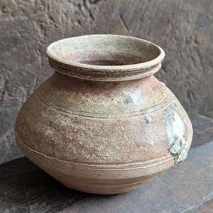 May include: A small, antique-style ceramic pot with a rounded body and a slightly flared rim. The pot is primarily a muted brown color with lighter tan and beige accents. Horizontal lines encircle the body, and a small patch of green is visible on the side.