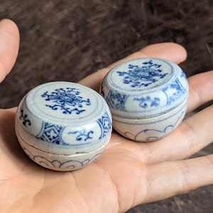 Op de afbeelding: Twee kleine, ronde keramische potjes met deksel, voorzien van een witte basis met ingewikkelde blauwe bloem- en geometrische patronen. Elke pot heeft een craquelé glazuurafwerking en een bijpassend deksel met gedetailleerde blauwe motieven.