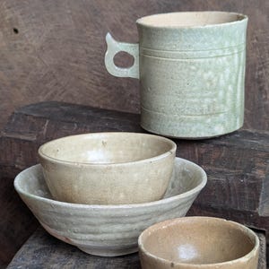 May include: A collection of handmade ceramic tableware in muted tones. The set includes a tall mug with a unique handle, two bowls, and a small, footed bowl. The pieces have a rustic, textured appearance, suggesting a natural, earthy aesthetic.