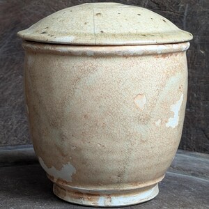 May include: An antique, cream-coloured ceramic jar with a lid. The jar has a rounded body and a flared base. The surface shows signs of age with a crackled glaze and areas of discolouration, giving it a rustic appearance.