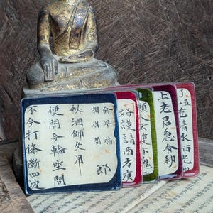 May include: A seated Buddha statue with a gold and dark finish positioned behind a row of five rectangular cards. Each card is encased in a clear plastic sleeve and features black Chinese calligraphy on a cream background with colored borders.