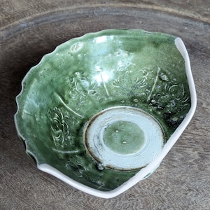 May include: A green ceramic bowl with a white rim and a raised design. The bowl is slightly angled and has a textured surface.
