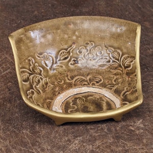May include: A small, square-shaped ceramic dish with a green glaze and intricate floral designs. The dish has three small feet and a slightly raised rim.