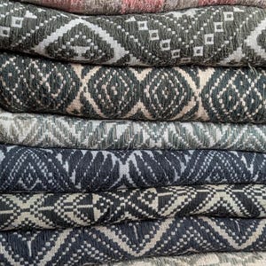 May include: A pile of woven textiles in various colours and patterns. The top textile is a light beige with a pink geometric design. Below are textiles in shades of green, grey, and blue, each with a repeating geometric pattern in white.
