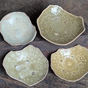 May include: Four handmade ceramic bowls in shades of green and tan. Each bowl has a unique textured or patterned design, including floral and honeycomb patterns. The bowls are arranged on a dark wooden surface.