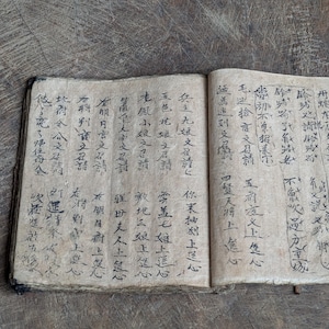 May include: An open antique book with aged, textured pages. The pages are filled with black calligraphic text. The book is lying on a weathered wooden surface, creating a vintage aesthetic.