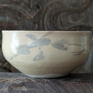 Könnte beinhalten: Eine cremefarbene Keramikschale mit einer glänzenden, rissigen Glasuroberfläche. Die Außenseite zeigt ein handgemaltes, abstraktes graues Blumenmuster mit feinen Pinselstrichen. Die Schale hat eine abgerundete Form mit einem glatten Rand.