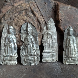 May include: Four silver-colored, antique religious figures. Each figure is in a standing pose, with detailed clothing and headwear. The figures are set against a rough, dark brown background, suggesting an aged appearance. The figures are likely made of metal.