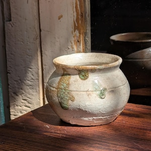 May include: A small, off-white ceramic pot with a rounded body and a wide opening. The pot has a textured surface with green and brown accents. Another similar pot is visible in the background, reflecting in the window.