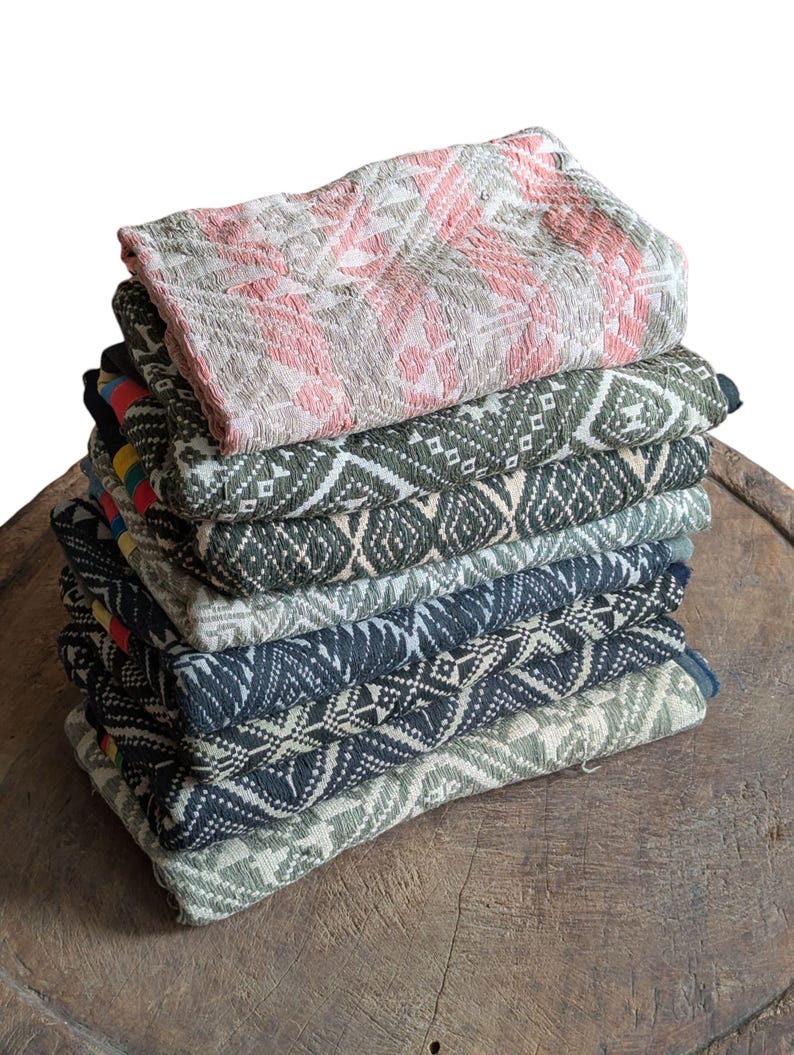 May include: A pile of folded textiles with geometric designs in various colours. The top textile is pink and grey, while the others feature shades of green, blue, and grey. The textiles are stacked on a wooden surface.