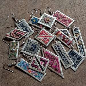 Op de afbeelding: Een verzameling van 12 paar zilveren oorbellen met kleurrijke stoffen accenten. De oorbellen zijn allemaal in verschillende vormen en maten en zijn in een willekeurig patroon gerangschikt. De stoffen accenten zijn allemaal in verschillende kleuren en patronen en zijn allemaal zeer ingewikkeld.