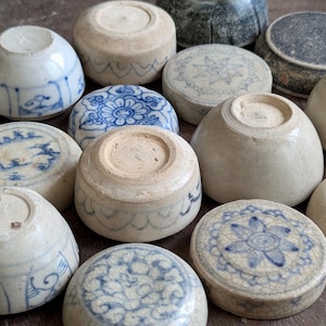 Può includere: Una collezione di piccole ciotole e coperchi in ceramica rotondi, alcuni con motivi floreali o geometrici blu e bianchi, altri in bianco sporco o grigio scuro. Gli oggetti mostrano segni visibili di usura, incluse crepe superficiali.