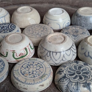 Könnte beinhalten: Eine Sammlung kleiner, runder Keramikschalen, die umgedreht präsentiert werden. Die Schalen weisen verschiedene handbemalte blaue, grüne und graue Muster auf, darunter florale Motive, geometrische Linien und Blütenblattdesigns auf cremefarbenen Oberflächen.