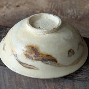 May include: A small, shallow ceramic bowl with a cream-colored glaze and irregular, dark brown, brush-like markings around the exterior. The bowl features a circular foot ring at the base and a smooth, rounded rim, showcasing a hand-crafted appearance.