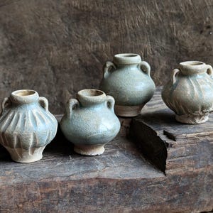 May include: Four small, glazed ceramic pots with handles. The pots are a light blue-green color and have a rustic, aged appearance. They are arranged on a wooden surface.