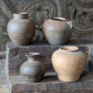May include: A collection of four small, weathered ceramic vessels in varying shades of brown and grey. The pots feature rounded bodies, short necks, and textured, earthy surfaces with visible signs of age, including subtle speckling and horizontal lines.