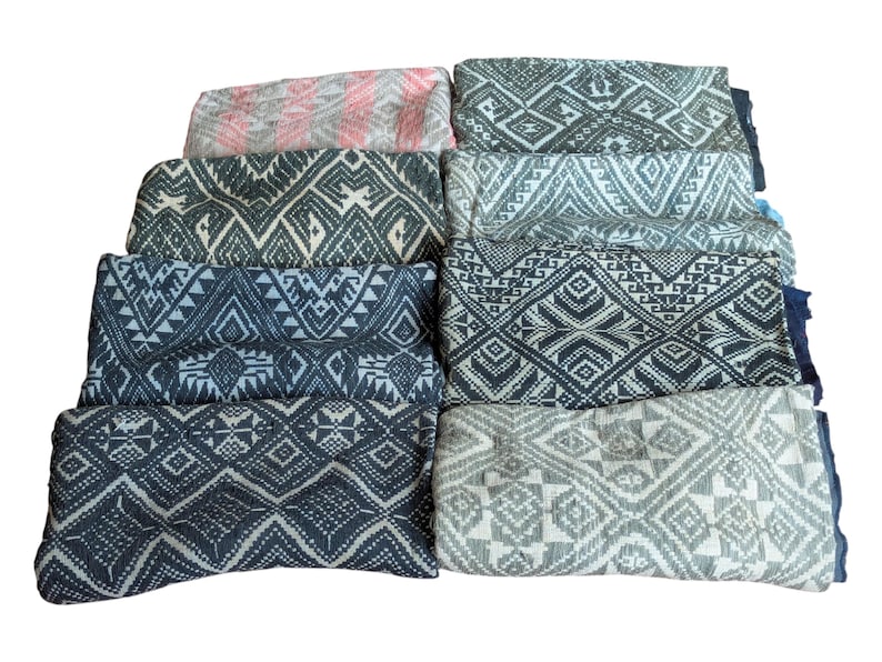 May include: A collection of folded fabric scarves with geometric patterns in various colours. The scarves feature designs in shades of grey, blue, and pink. The patterns include diamonds, triangles, and other shapes.