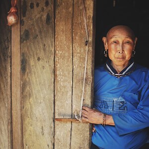 May include: A woman with short, shaved hair stands in a doorway. She is wearing a blue shirt with a white collar and a silver necklace. She has large silver earrings and a serious expression on her face.