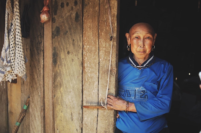 May include: A woman with short, shaved hair stands in a doorway. She is wearing a blue shirt with a white collar and a silver necklace. She has large silver earrings and a serious expression on her face.