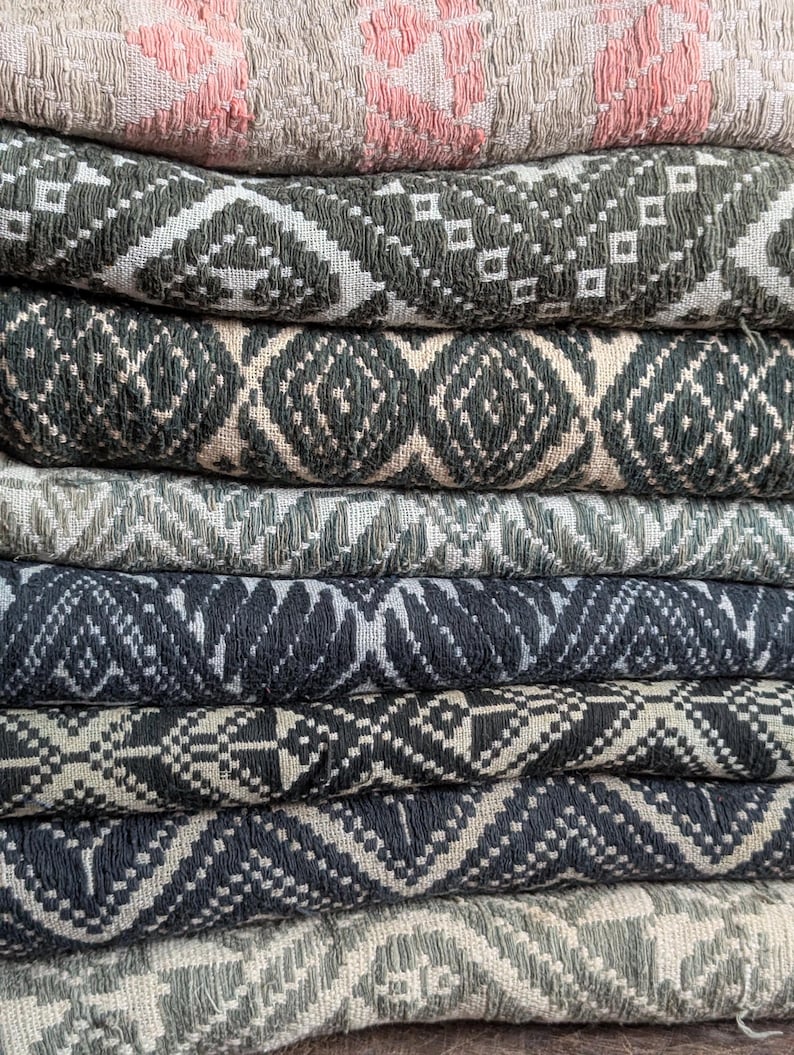May include: A pile of woven textiles in various colours and patterns. The top textile is a light beige with a pink geometric design. Below are textiles in shades of green, grey, and blue, each with a repeating geometric pattern in white.