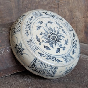 May include: A round, antique ceramic container with a lid. The container is off-white with blue floral and geometric patterns. The lid features a central floral design and is surrounded by intricate detailing. The container is resting on a wooden surface.