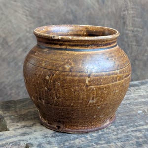 May include: A small, round, brown ceramic pot with a wide opening. The pot has a textured surface with horizontal lines and a dark band near the rim. The pot is sitting on a wooden surface.