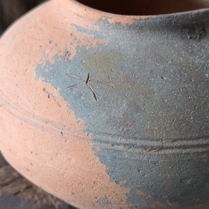 Puede incluir: Primer plano de una olla de terracota hecha a mano con un cuerpo redondeado y una abertura ancha. La olla presenta una combinación de colores de dos tonos, con una base naranja claro y una sección superior gris-azul más oscura y desgastada. Un diseño en forma de estrella está grabado.