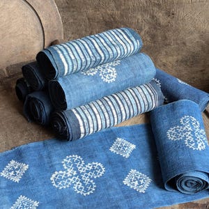 May include: A stack of indigo blue fabric table runners with white embroidered designs. The runners are folded and stacked on a wooden surface.
