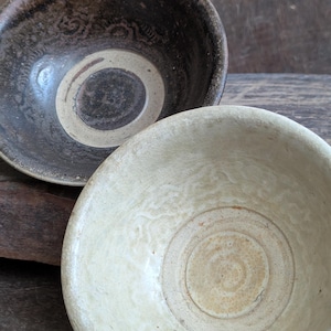 May include: Two ceramic bowls. One is dark brown with a cream-coloured ring and a darker centre. The other bowl is cream-coloured with a concentric circle design in the centre. The bowls are set against a dark wooden surface.