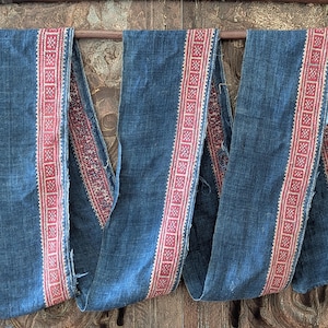 May include: A long strip of dark blue fabric featuring a woven red and white geometric border pattern. The textile has a visible coarse weave and frayed edges, draped over a horizontal wooden rod. The design consists of repeating square motifs.