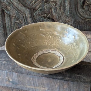 Puede incluir: Un cuenco de cerámica redondo con un esmalte de color verde oliva apagado y acabado brillante. El interior presenta un patrón floral en relieve en los laterales y un diseño de anillo circular en la base, con un borde ligeramente irregular.