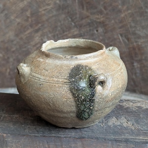 May include: A small, round, antique ceramic pot with two small handles. The pot is a mottled beige color with a dark green and black stain on the side. The rim is uneven, and the pot has a rustic, aged appearance.