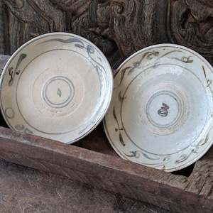 May include: Two antique, off-white ceramic plates with hand-painted gray and brown decorative patterns. The plates have a central design and a border of stylized leaves and swirls. The plates are displayed in a rustic wooden tray.