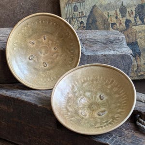 May include: Two small, round, ceramic bowls with a light brown glaze and a decorative pattern. The bowls are sitting on a wooden surface.