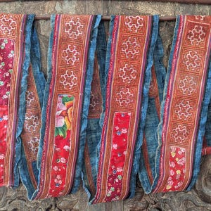 Könnte beinhalten: Ein langer, mehrlagiger Textilstreifen mit einem Patchwork-Design aus blauem, rotem und orangefarbenem Stoff. Das Stück enthält komplexe geometrische Stickmuster, blumig bedruckte Stoffbahnen und ausgefranste Kanten, drapiert über einer Holzstange.