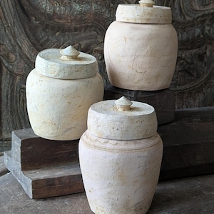 May include: Three off-white ceramic jars with lids. The jars have a rounded shape and are topped with a flat lid and a small conical knob. The jars are arranged on a wooden surface, with a dark, carved wooden background.