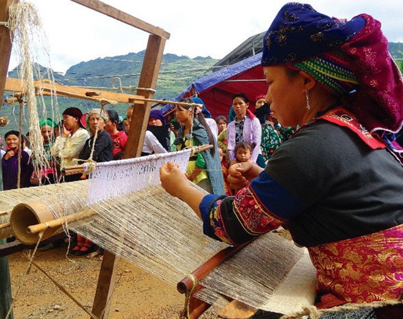 May include: A woman in traditional dress weaving on a wooden loom. The loom is set up outdoors in front of a crowd. The woman wears a dark blue head covering with red and pink accents.
