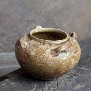 Pode incluir: Pequeno pote redondo de cerâmica antiga com uma abertura larga e duas pequenas alças. O pote tem uma superfície texturizada e rachada em tons de castanho e bege, com um interior mais escuro. Provavelmente para fins decorativos.